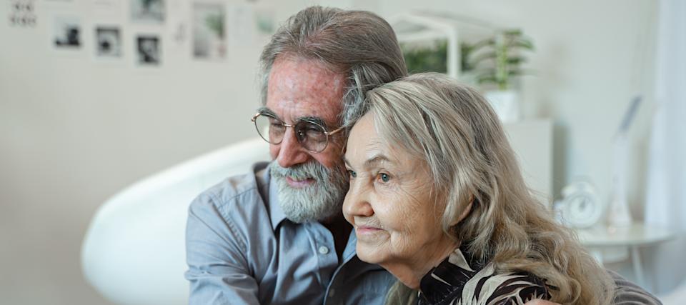 An older couple hold each other closely and look into the distance.