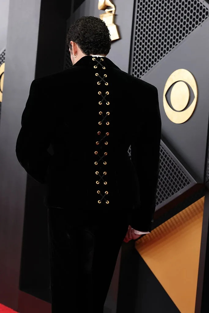 Person on red carpet wearing a velvet suit with eyelet lace-up detail on the back. Geometric background includes a notable award logo