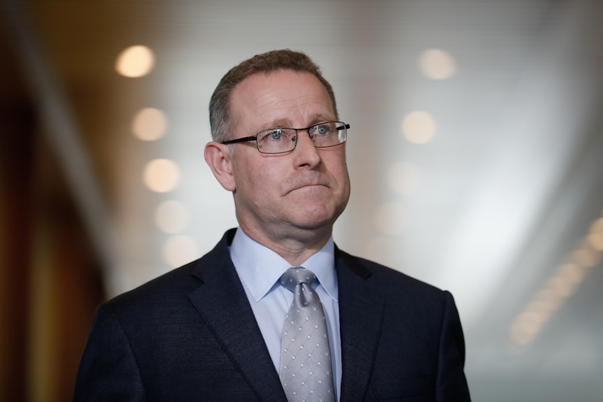a man wearing glasses standing indoors talking to the media