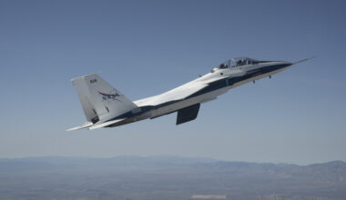 A white and blue NASA F-15 research jet climbs to altitude with an approximately 3-foot experimental wing design mounted beneath its fuselage. Viewed in profile against a blue sky with mountains in the distance, the test article resembles a ventral fin below the aircraft.
