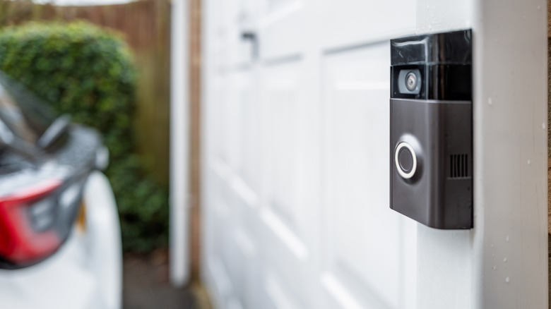 Shallow focus of a popular, battery powered wireless doorbell and integral camera seen located on a driveway, near a side gate entrance. A parked car is on the driveway.