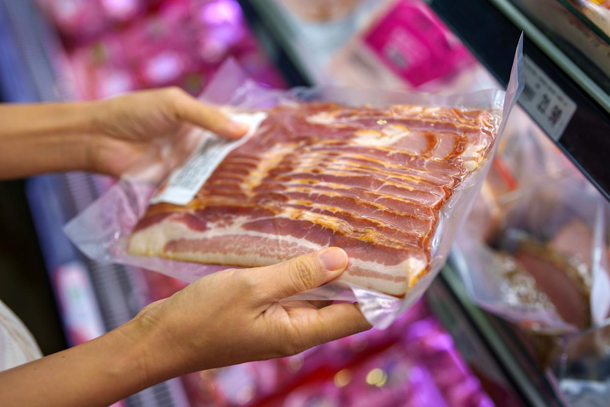 A cropped shot of a woman carefully selecting raw pork bacon in the processed meat section of a supermarket.
