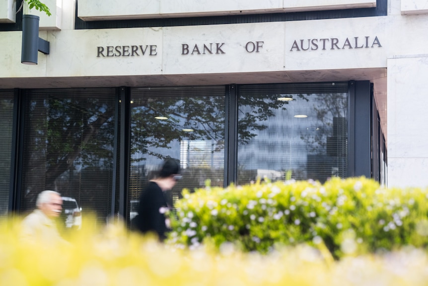 People walk past the exterior of the Reserve Bank building in Canberra, which has a hedge in front of it.
