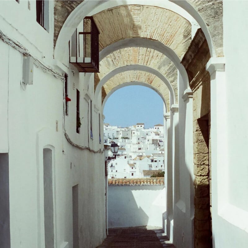A narrow alleyway with white walls and stone arches leads to a view of a sunlit town with white buildings, set against a clear blue sky. Shadows and light create a serene, Mediterranean atmosphere.