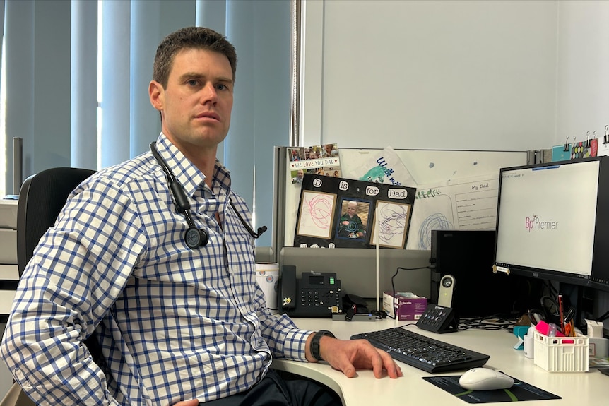 A man has short blonde hair, he sits wearing a checked top at a computer in a medical office with a stethoscope around neck