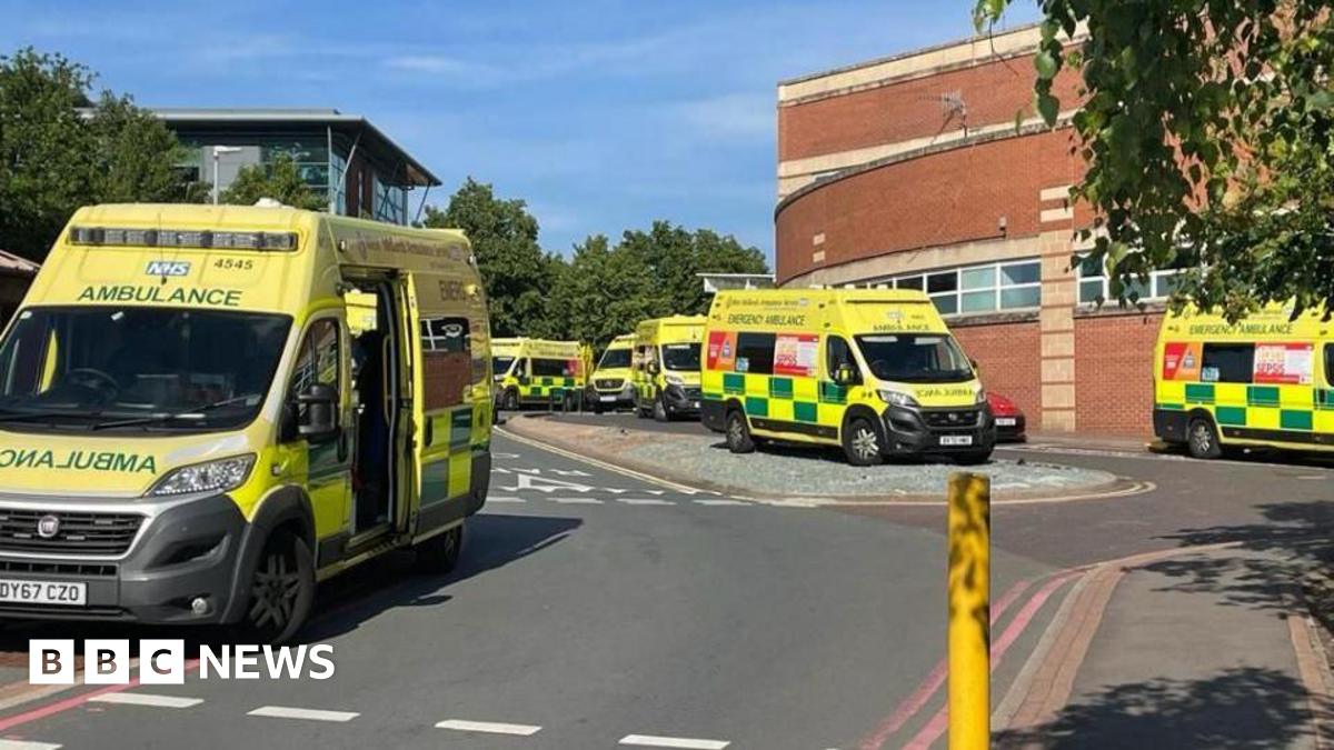 Six ambulances outside a hospital building. The vehicles are all mainly yellow with green rectangle patterns on. The hospital building is red brick and the lower part has a curved shape.