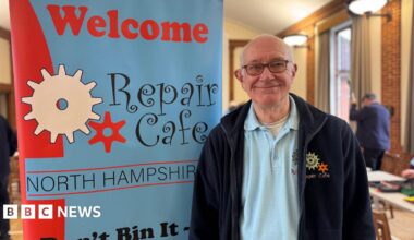 A man with white hair and glasses, wearing a navy fleece and blue polo shirt, both embroidered with a "repair cafe" logo. He is stood next to a blue and red banner which reads "welcome, repair cafe North Hampshire".