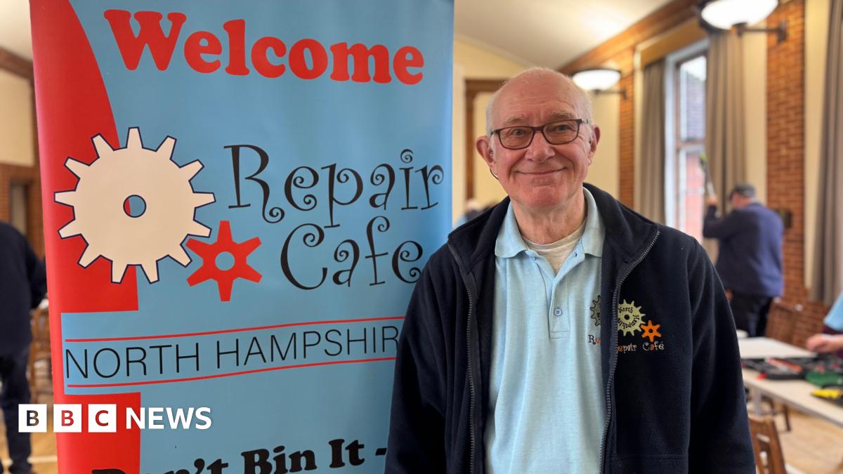 A man with white hair and glasses, wearing a navy fleece and blue polo shirt, both embroidered with a "repair cafe" logo. He is stood next to a blue and red banner which reads "welcome, repair cafe North Hampshire".
