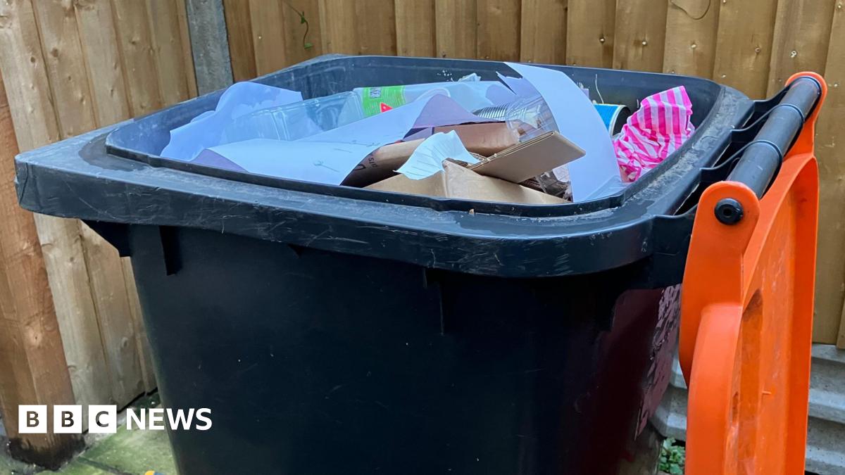 Dark grey wheelie bin with the orange lid opened, and paper and cardboard visible inside. The bin is in front of a wooden fence.