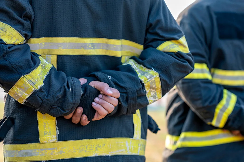 Firefighters in protective gear with reflective stripes stand in a line, hands behind their backs, appearing focused and ready