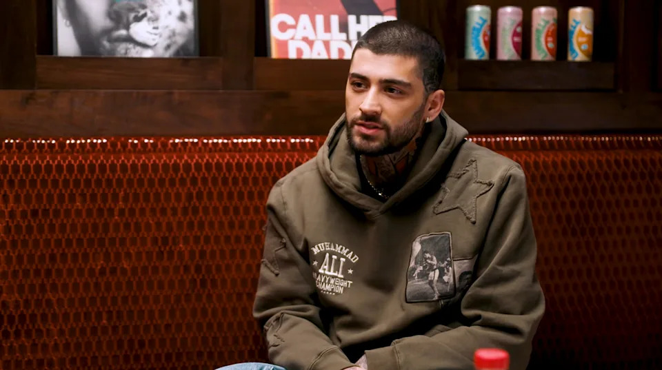 A person is seated in a room, wearing a casual hooded sweatshirt with graphic patches, next to shelves with posters and beverages