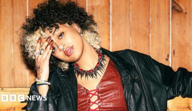 Nxdia with blond and black hair rests her head to the side on her hand. She is wearing a red leather-look top with a leather jacket and black and red choker necklace with spikes.