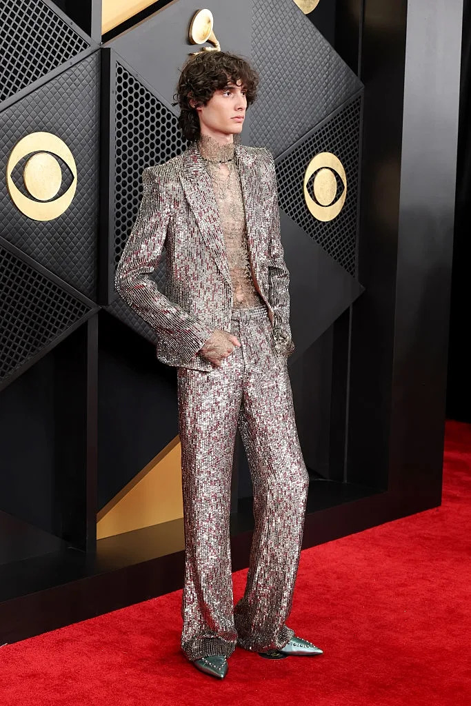 Person on red carpet wearing a glittering silver suit with a sheer shirt, posing in front of a geometric backdrop
