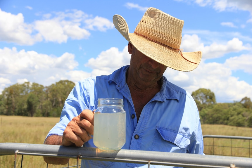 farmer in wide brimed hat and blue shirt holding a glass with cloudy water inside