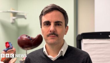 Tom Dolphin, a man with brown hair and a moustache, wears a grey jumper and a white shirt.