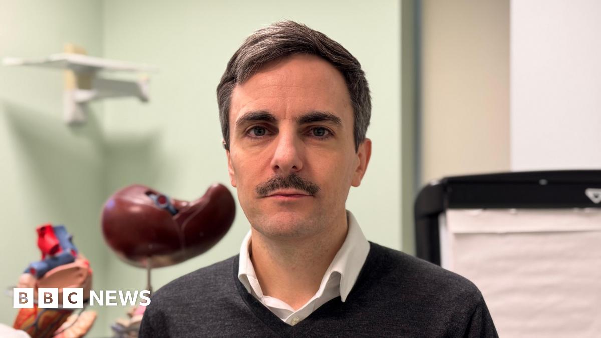 Tom Dolphin, a man with brown hair and a moustache, wears a grey jumper and a white shirt.