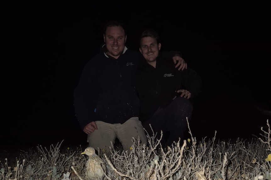 Two men crouch close to the ground. A small brown and white bird is in front of them.
