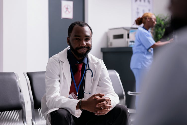 A man in a white coat with a stethoscope seated, with a woman in blue scrubs in the background