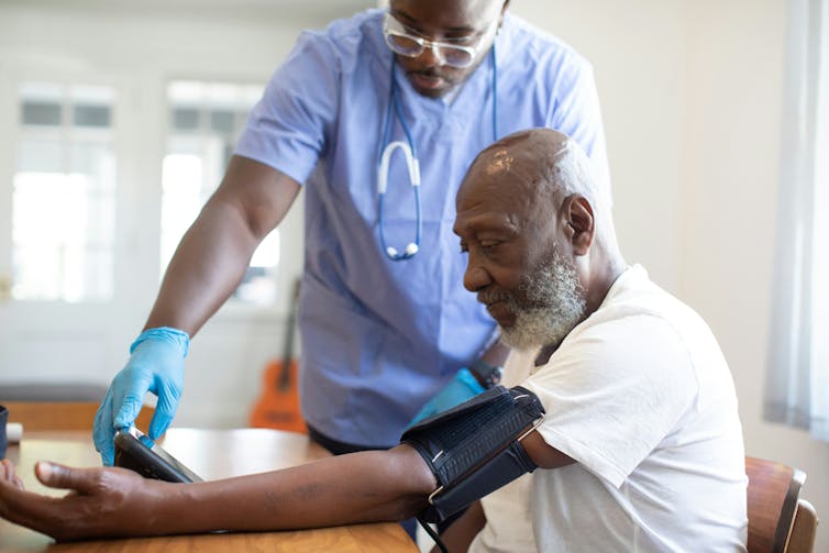 A man with a greying beard having his blood pressure taken by a health-care provider in blue scrubs