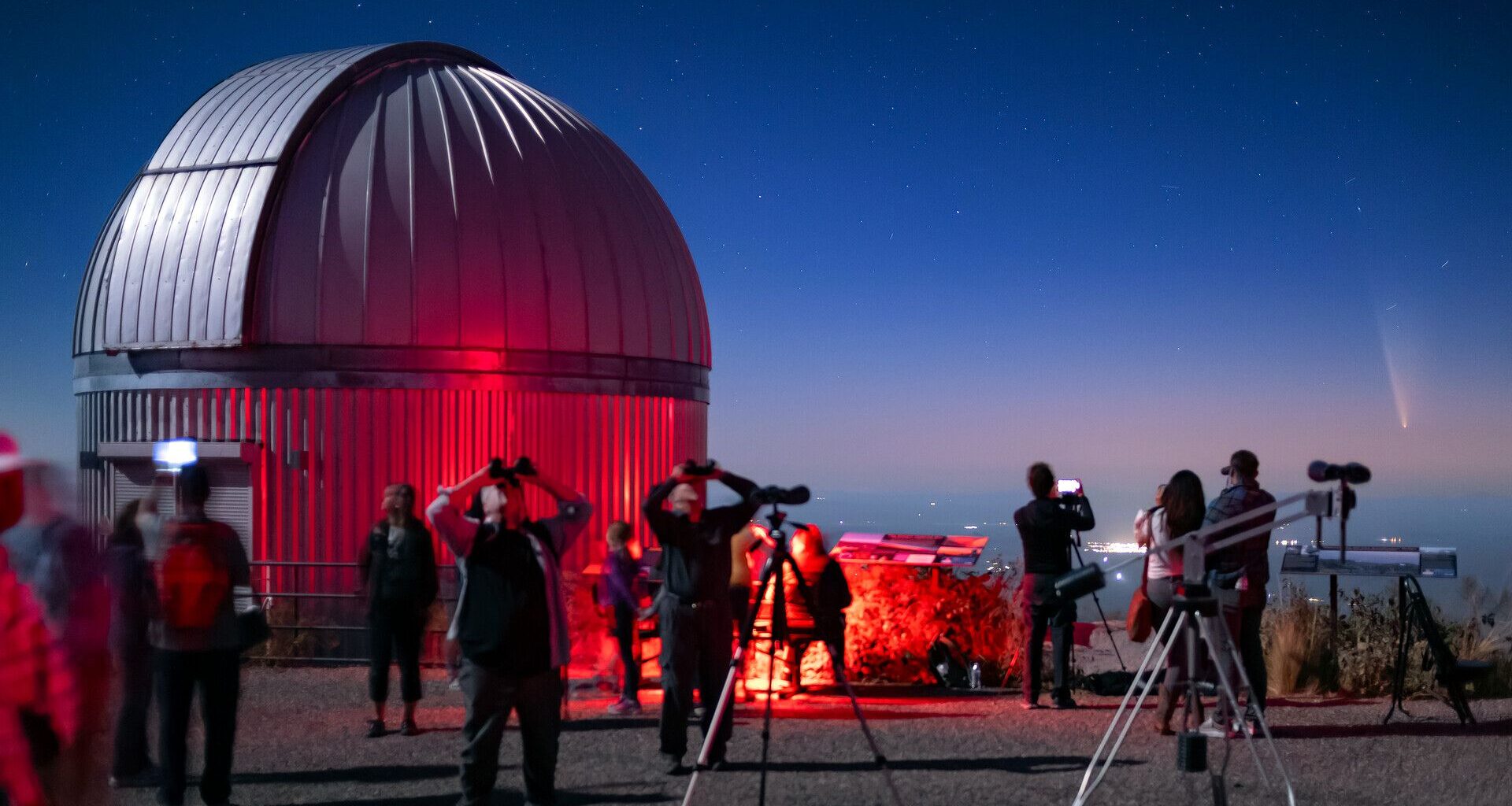 A circular dome of a telescope is illuminated in red light as a crowd of people holding binoculars and telescopes look at a pink and blue night sky.