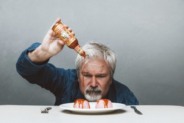 man with silver gray hair pouring tabasco sauce on a plate with two lumps of food on it