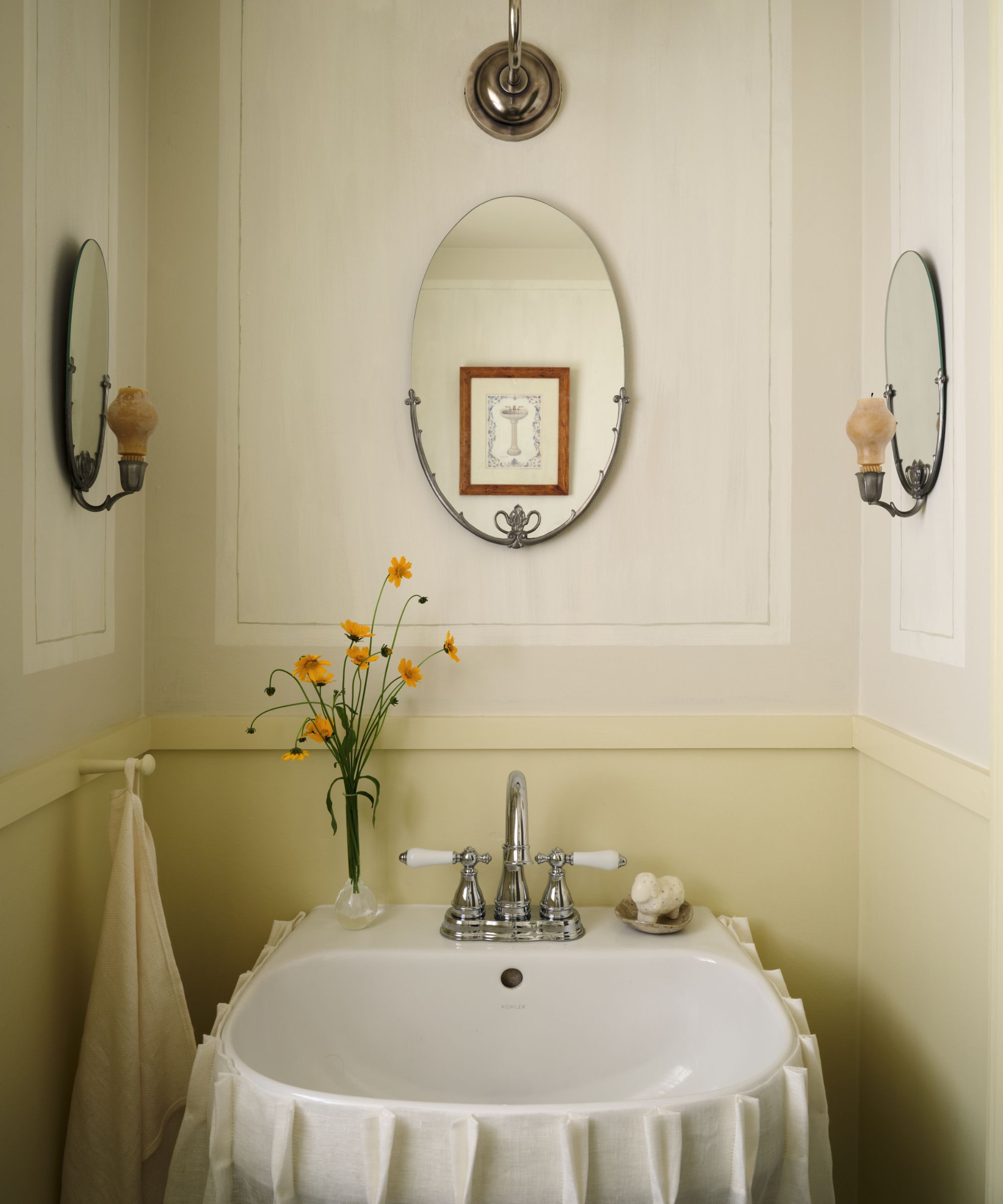 A powder room with butter yellow lower walls, panelled walls in a warm white on the upper walls, a white sink, and an oval wall mirror above it.