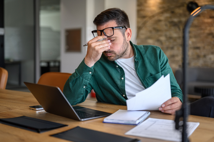 A man rubs his eyes while analyzing reports at his desk.