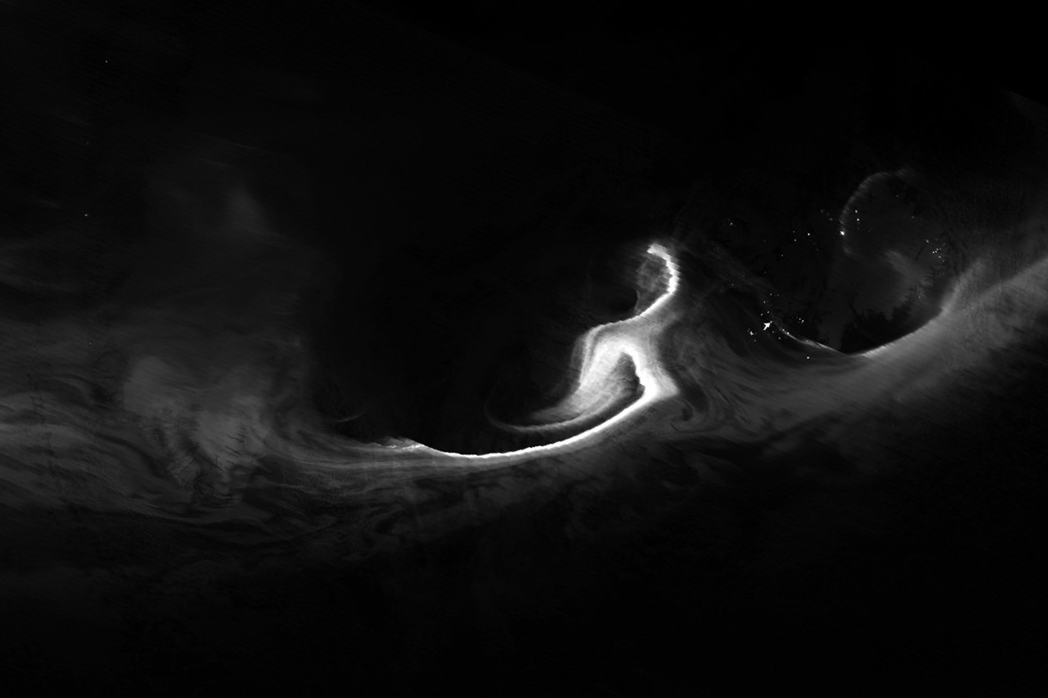 A nighttime satellite image shows a grayscale view of the northern lights over the Denmark Strait. Wisps of light stretch from Greenland to Iceland, with the brightest light just west of Iceland. Reykjavík city lights appear as a small dot.