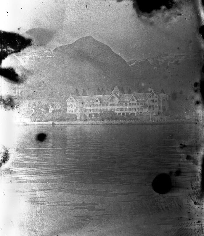 A faded, black-and-white photograph of a large, ornate building on the shore with a mountain in the background and water in the foreground. The image is damaged with dark stains and spots.