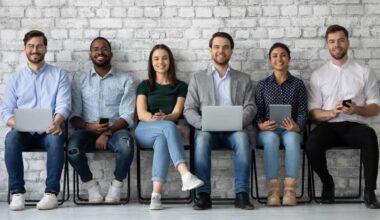 a line of job applicants sit on stools against a brick wall in an office environment, various holding laptops , devices and paper, as though waiting to be interviewed for a position.
