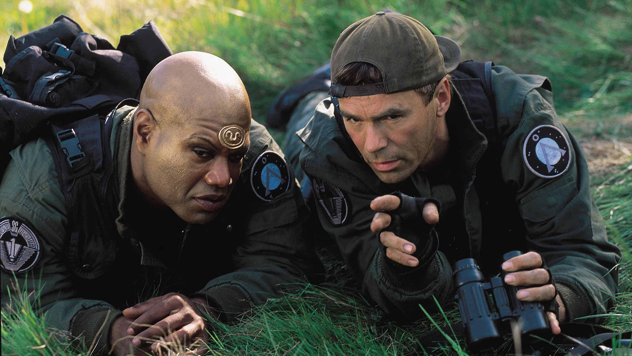 Two men in military gear are laying down prone on grass while talking to each other.