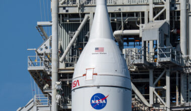 The Orion spacecraft is white and cylinder-shaped with a pointed nose at the top. It rests above the SLS rocket, part of which (the orange part) can just barely be seen at the bottom of the photo. Behind Orion and the SLS is a gray gantry with scaffolding and balconies.