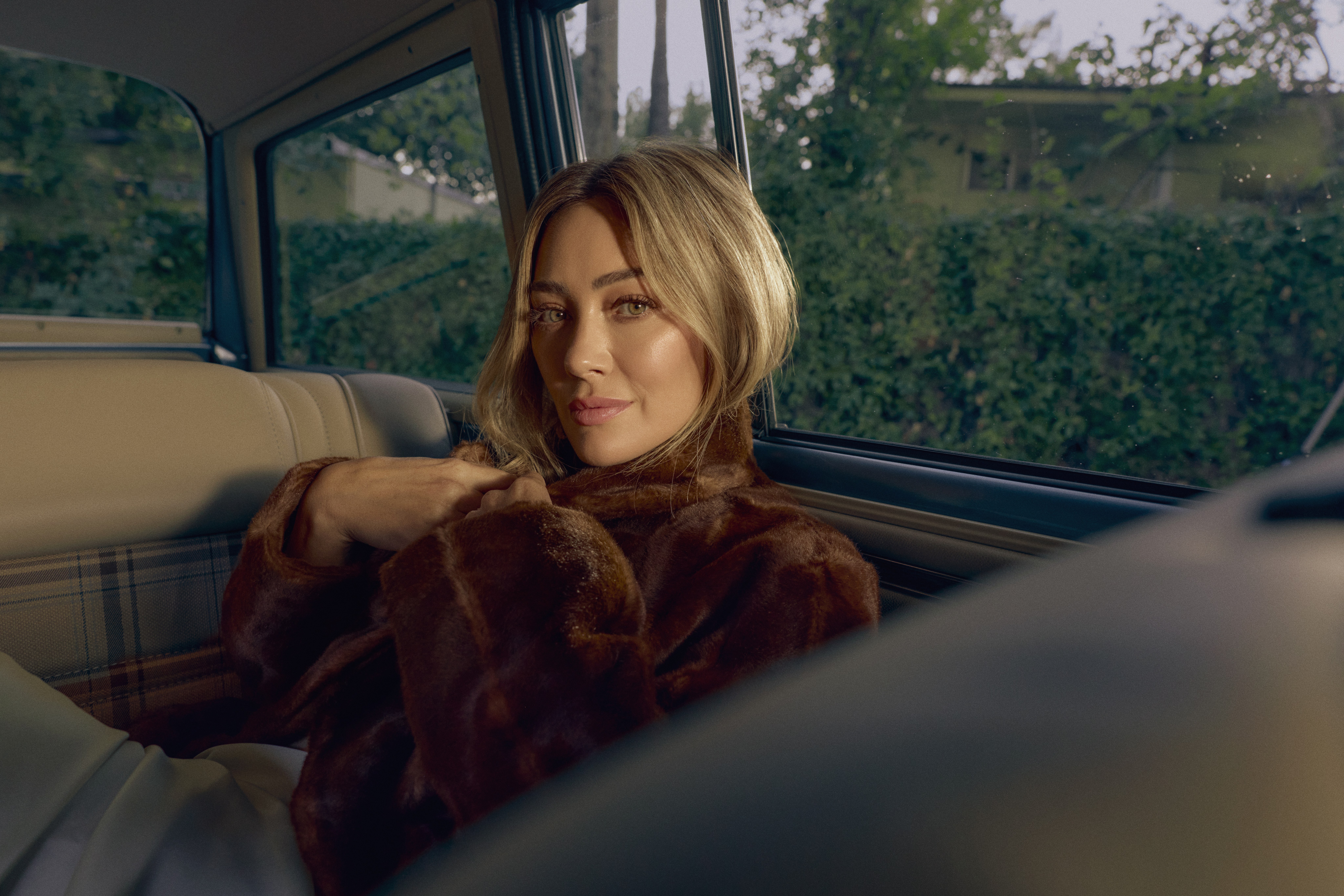 hilary duff wears a fur coat while lying in the back seat of a vintage car driving down a dimly lit neighborhood street in a photo promoting her album luck or something