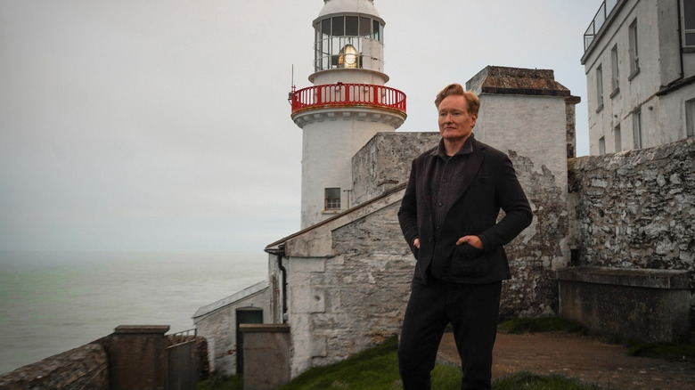 Conan O'Brien holds his hands in the pockets of his grey tweed suit while standing in front of a lighthouse in "Conan O'Brien Must Go"