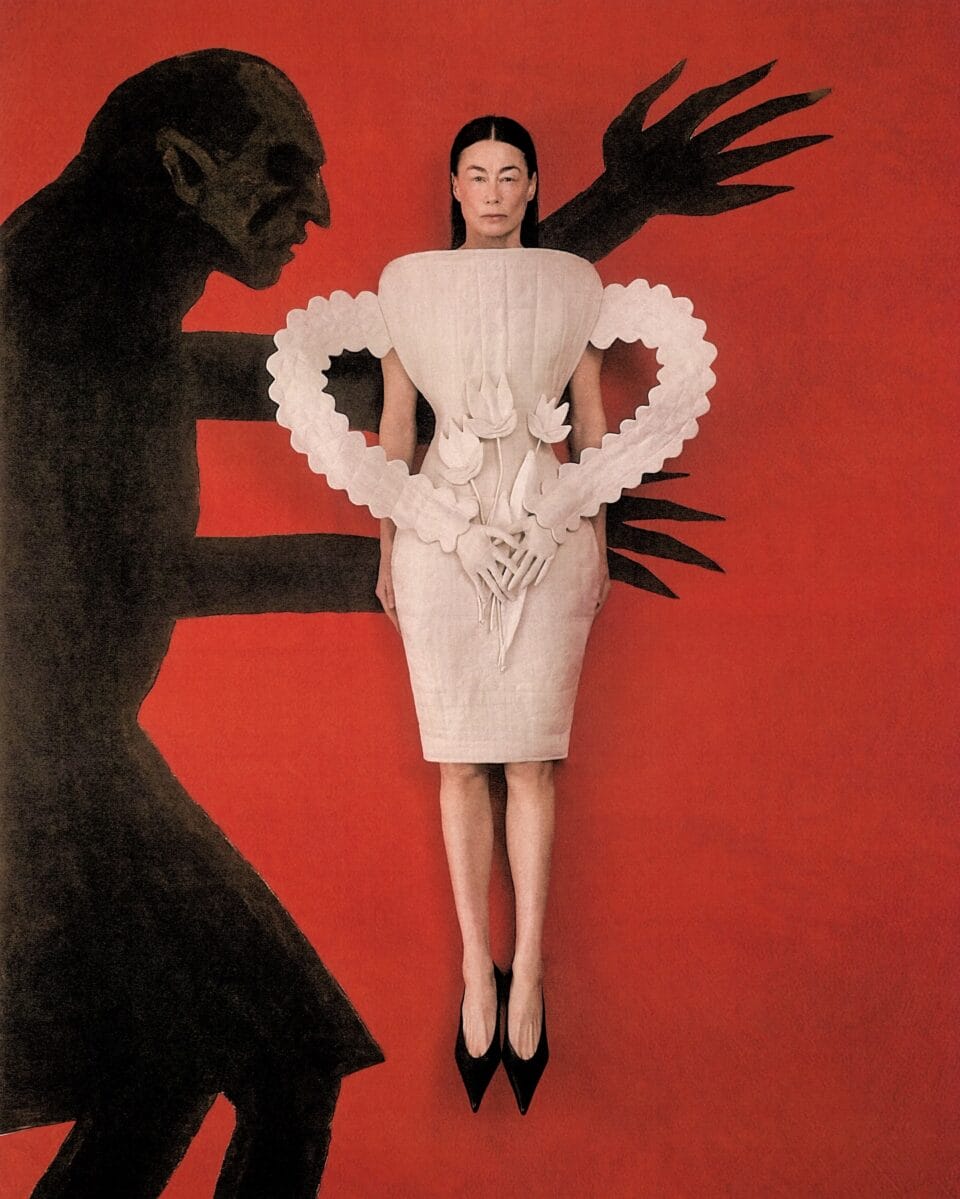 an editorial flat-lay portrait by Szilveszter Makó of a woman in a sculptural dress and pointy shoes positioned in front of the arms of a two dimensional silhouette of a man