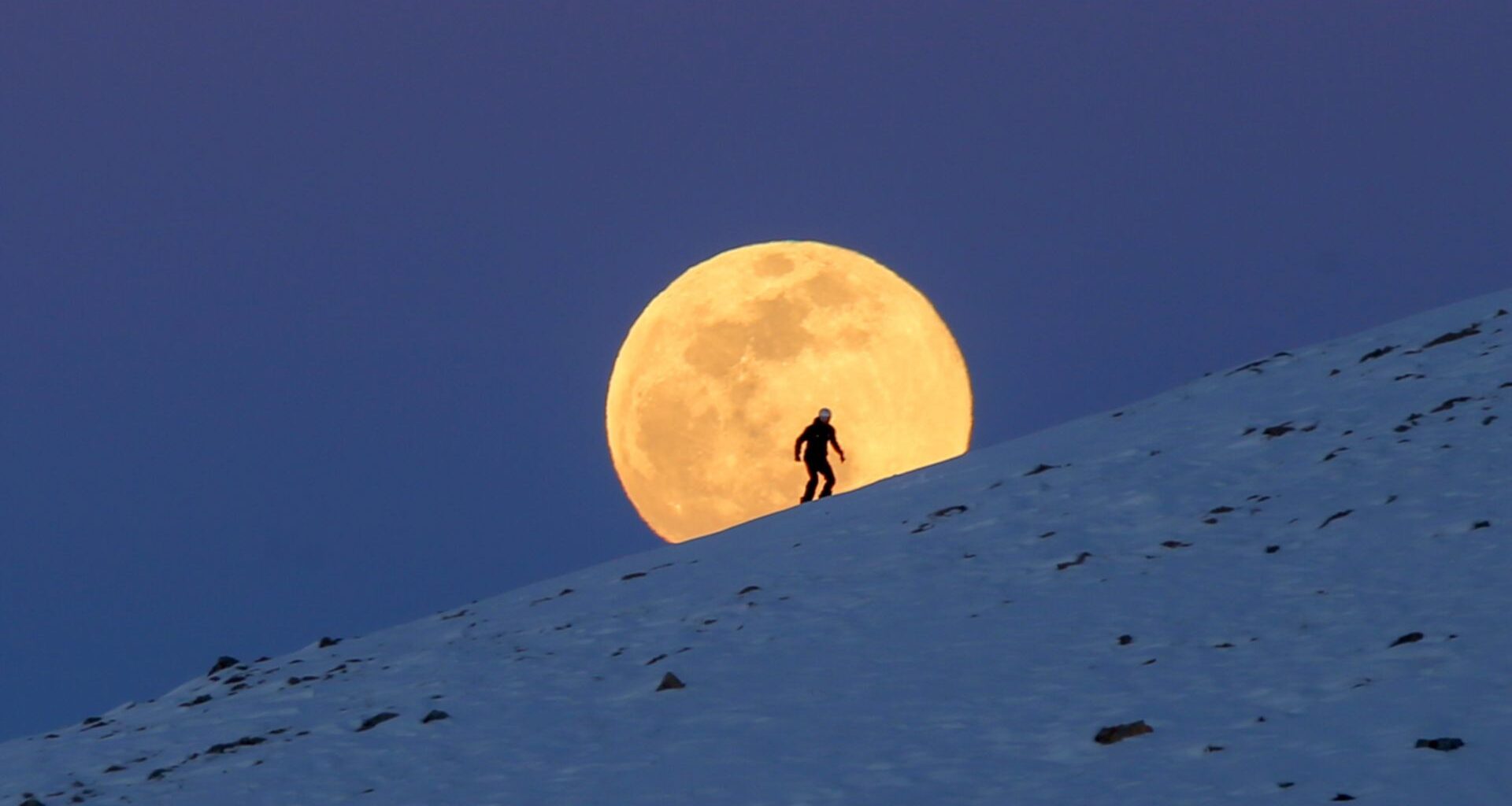 A yellow full moon is pictured rising over a snow covered mountain behind a skier in a darkening blue sky.
