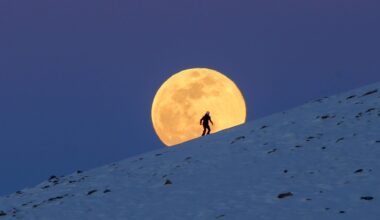 A yellow full moon is pictured rising over a snow covered mountain behind a skier in a darkening blue sky.