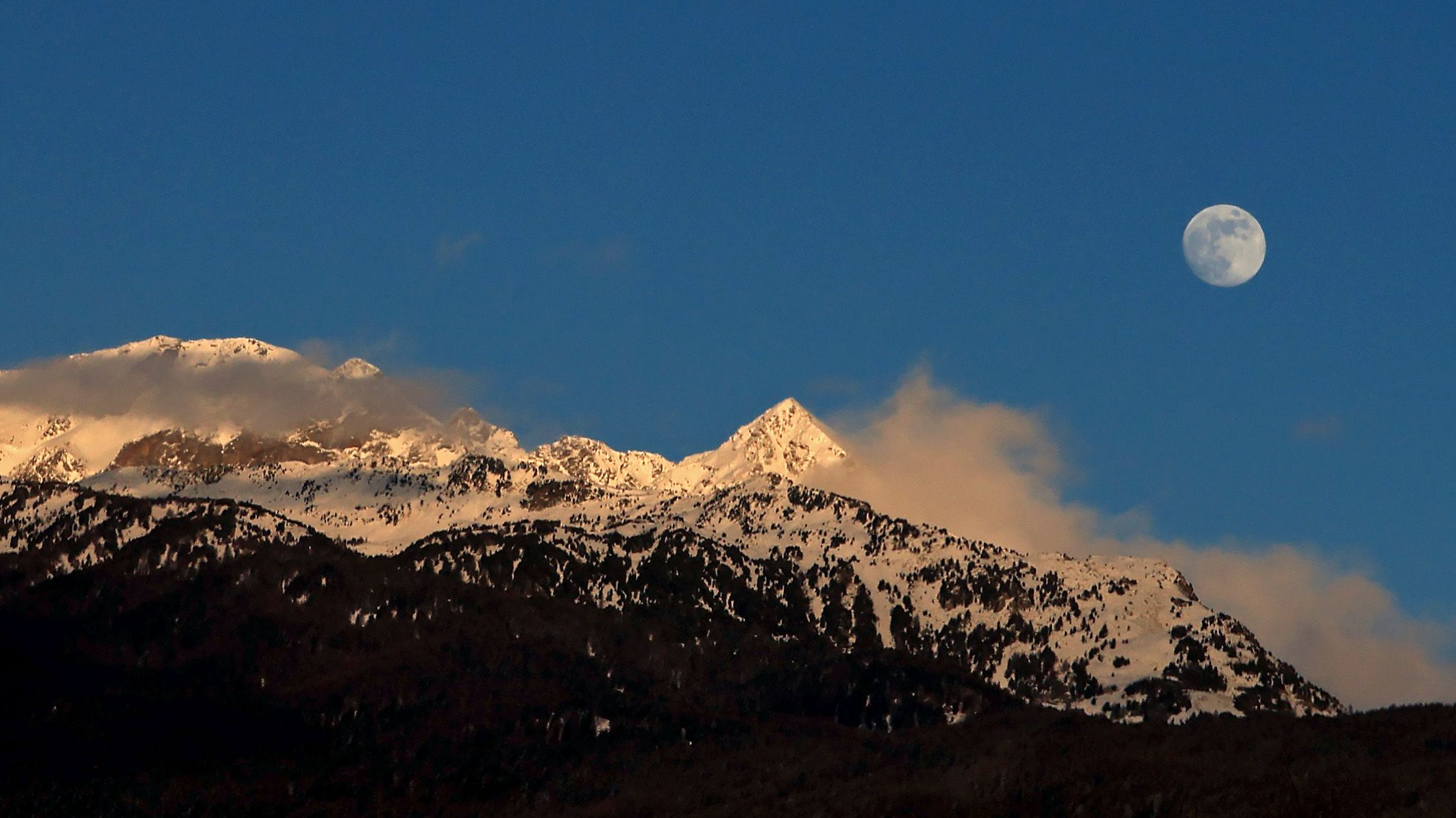 A full moon is pictured rising in a blue sky above snow capped mountains threaded with clouds.