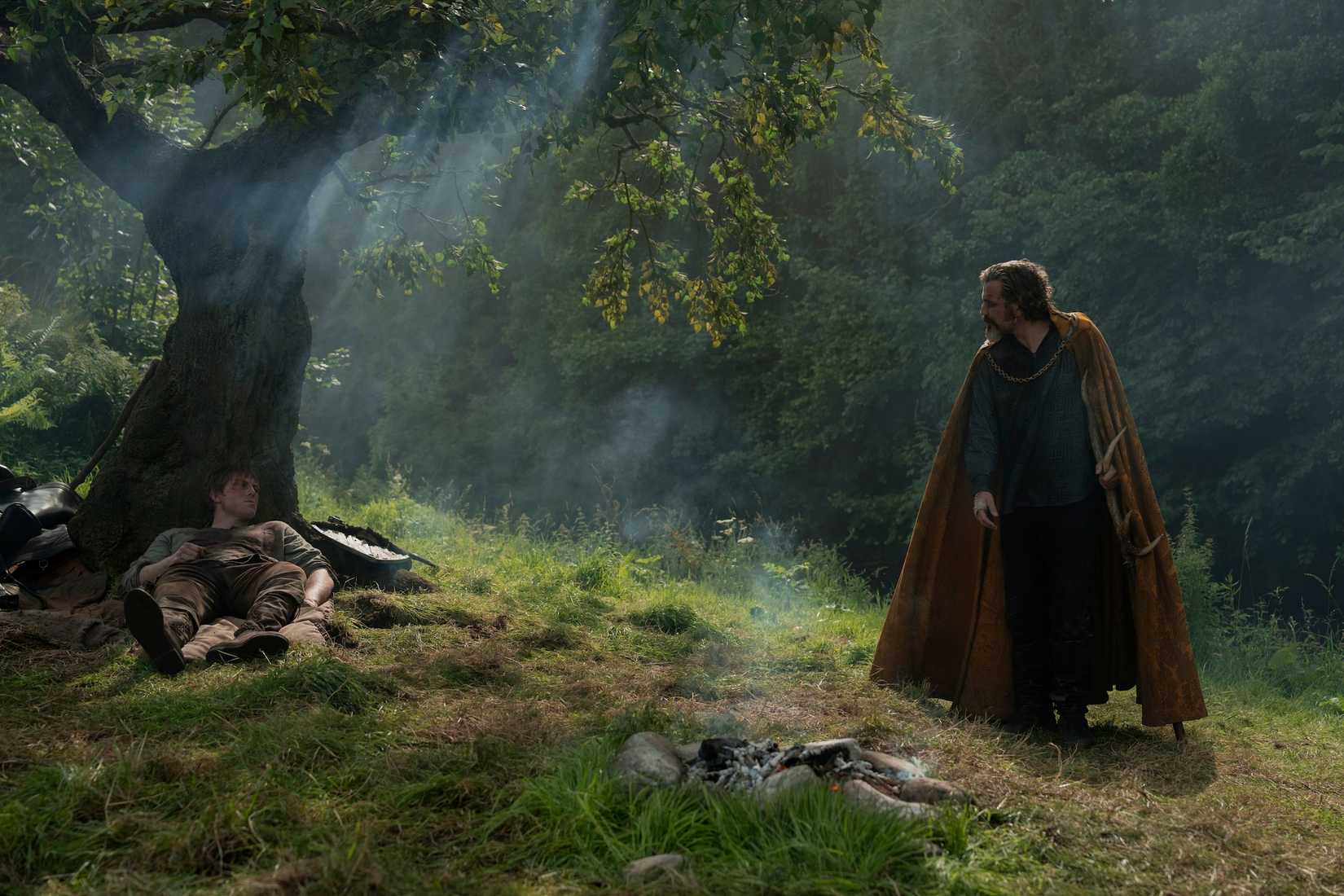 Dunk (Peter Claffey), a bruised, battered knight, lies under a tree while Ser Lyonel (Daniel Ings), a bearded knight in a yellow cloak, stands a distance away and shouts at him in A Knight of the Seven Kingdoms