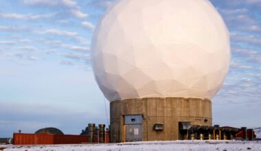 A white geodesic dome sits on top of a circular concrete base surrounded by snow under a cloudy blue sky