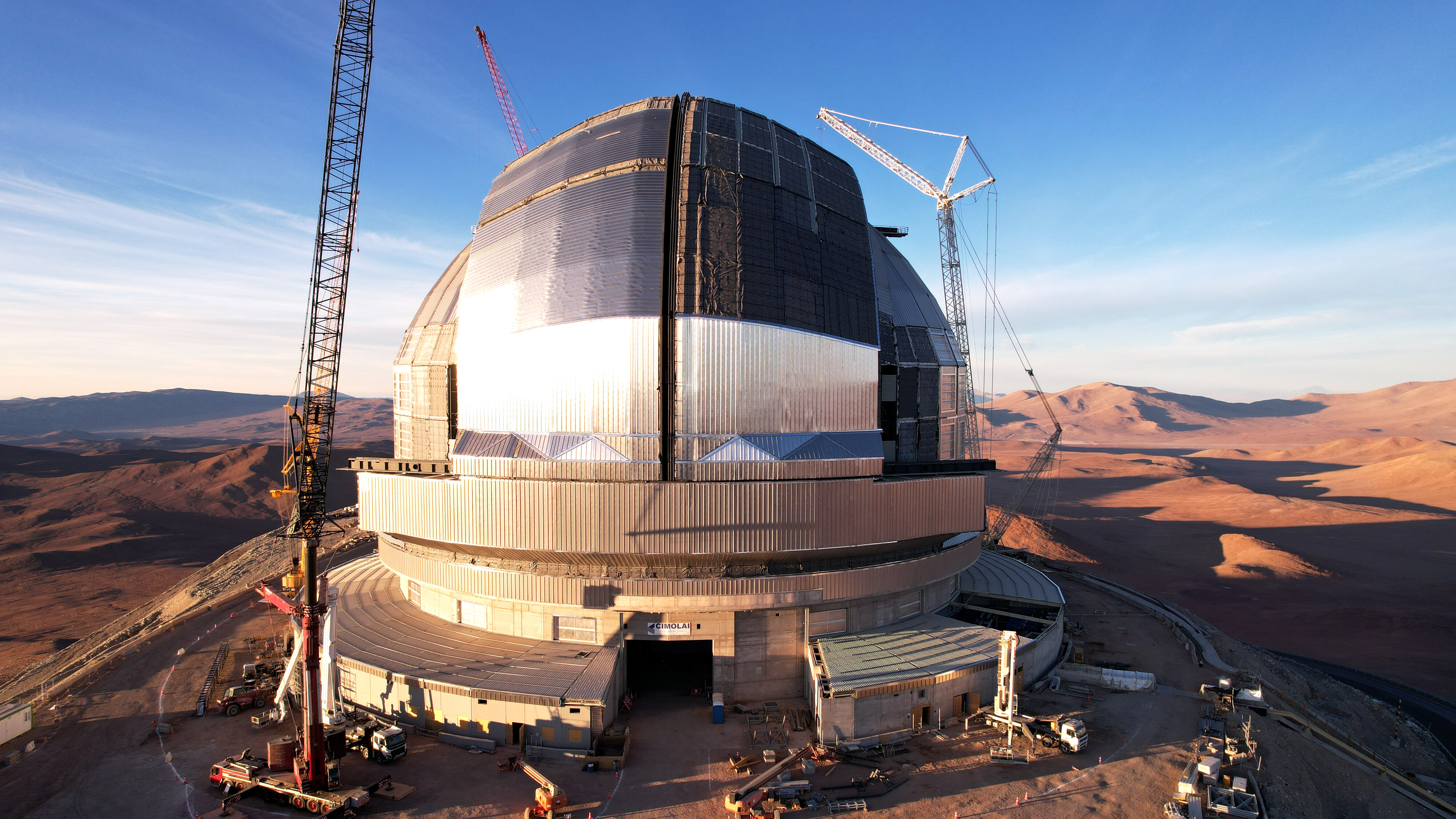 An enormous telescope dome is pictured in a desert during the day surrounded by cranes. Two huge silver sliding doors are visible, closed in the middle of the dome.