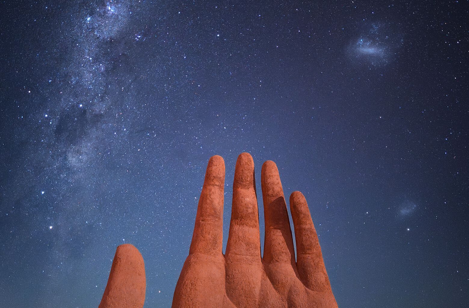 A large, surreal sculpture of a hand rises from the desert, under a starry night sky illuminated by the Milky Way
