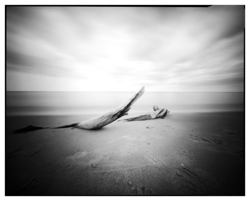A black and white photo of a beach with two large pieces of driftwood on the sand under a cloudy sky. The water and sky appear smooth and blurred, creating a serene, minimalist atmosphere.