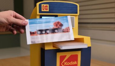 A hand holds a color photo in front of a small yellow and blue Kodak-branded photo printer on a wooden table, with window blinds in the background.
