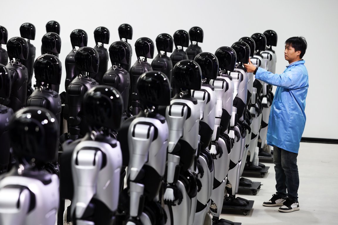 A man in a blue lab coat is inspecting or adjusting one of many humanoid AgiBot robots arranged in rows. The robots have black, glossy heads and silver and black bodies, standing on platforms with wheels. The background is plain white, emphasizing the robots and the man.