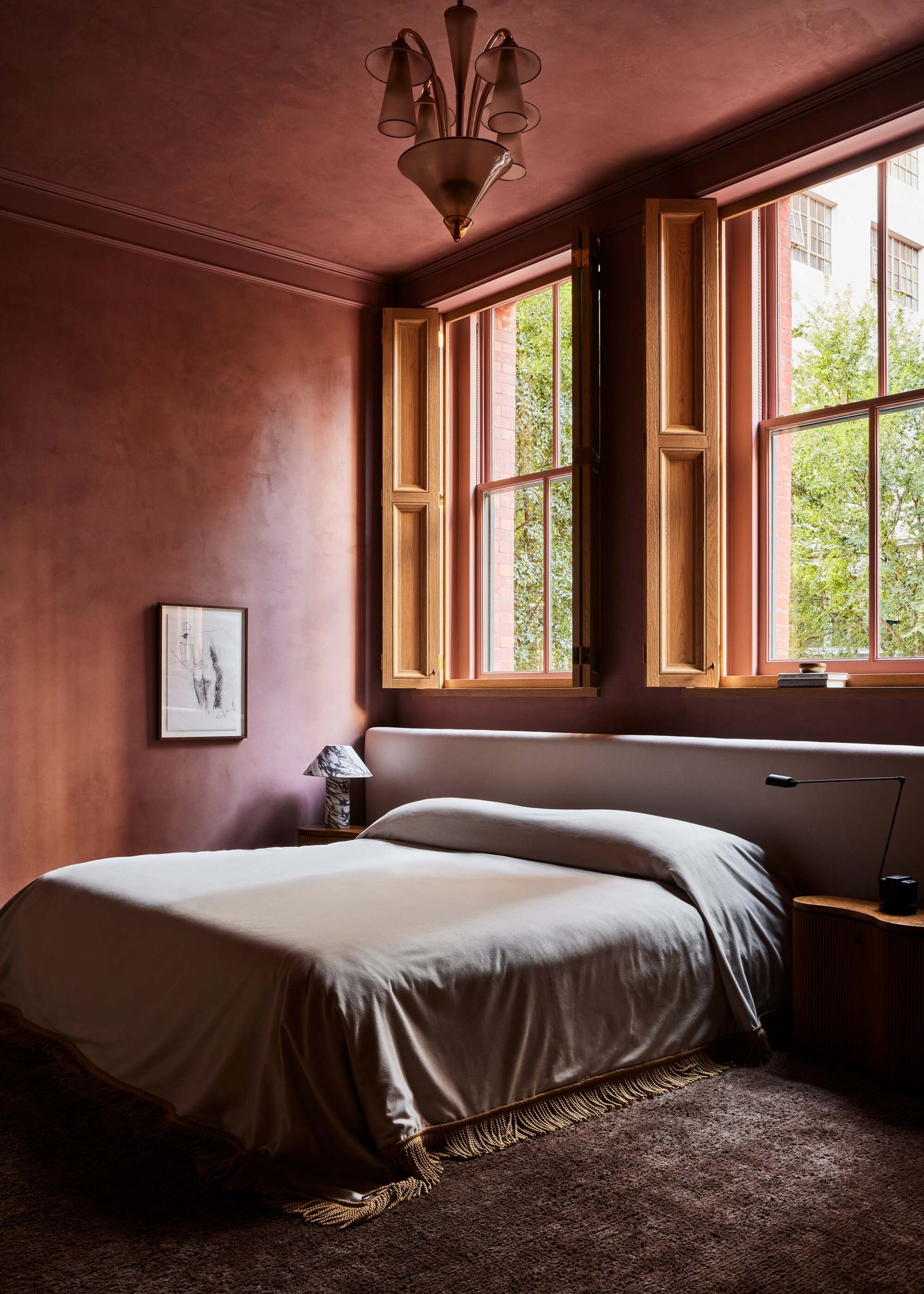color-drenched plastered walls in deep red, large windows, bedcover with fringe, brown rug, artwork on wall, and extended headboard