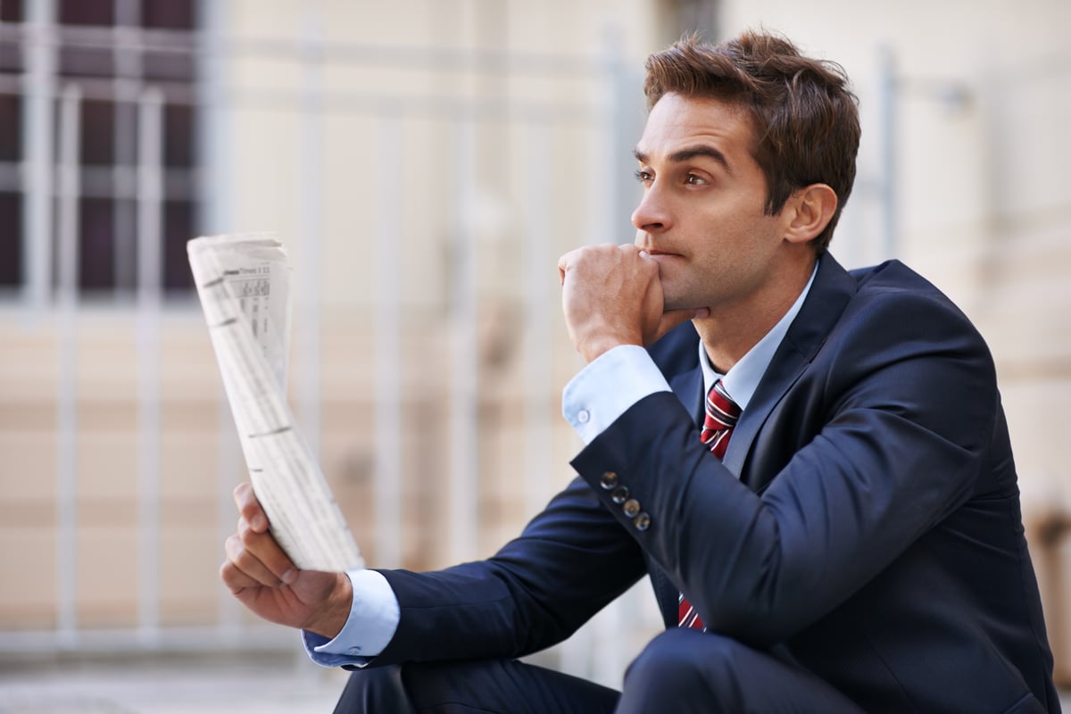 A well-dressed person holding a newspaper looks into the distance with a contemplative expression.