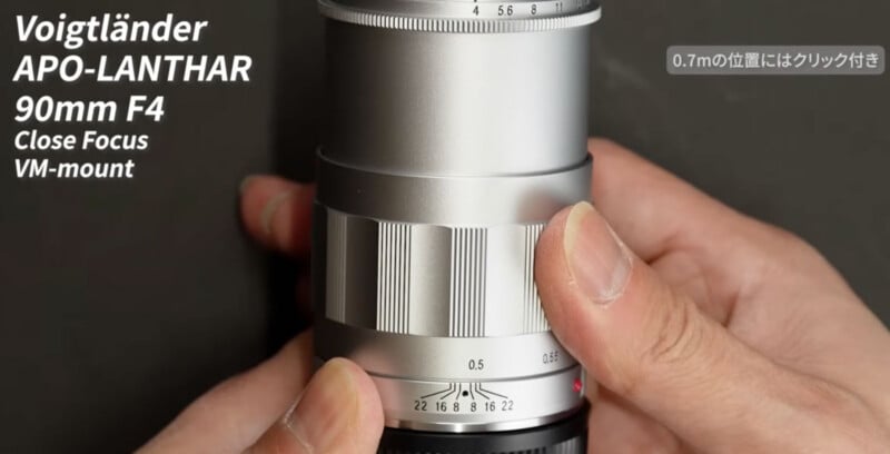 A person adjusts the focus ring on a silver Voigtländer APO-LANTHAR 90mm F4 Close Focus VM-mount camera lens, with product details displayed on the left side of the image.