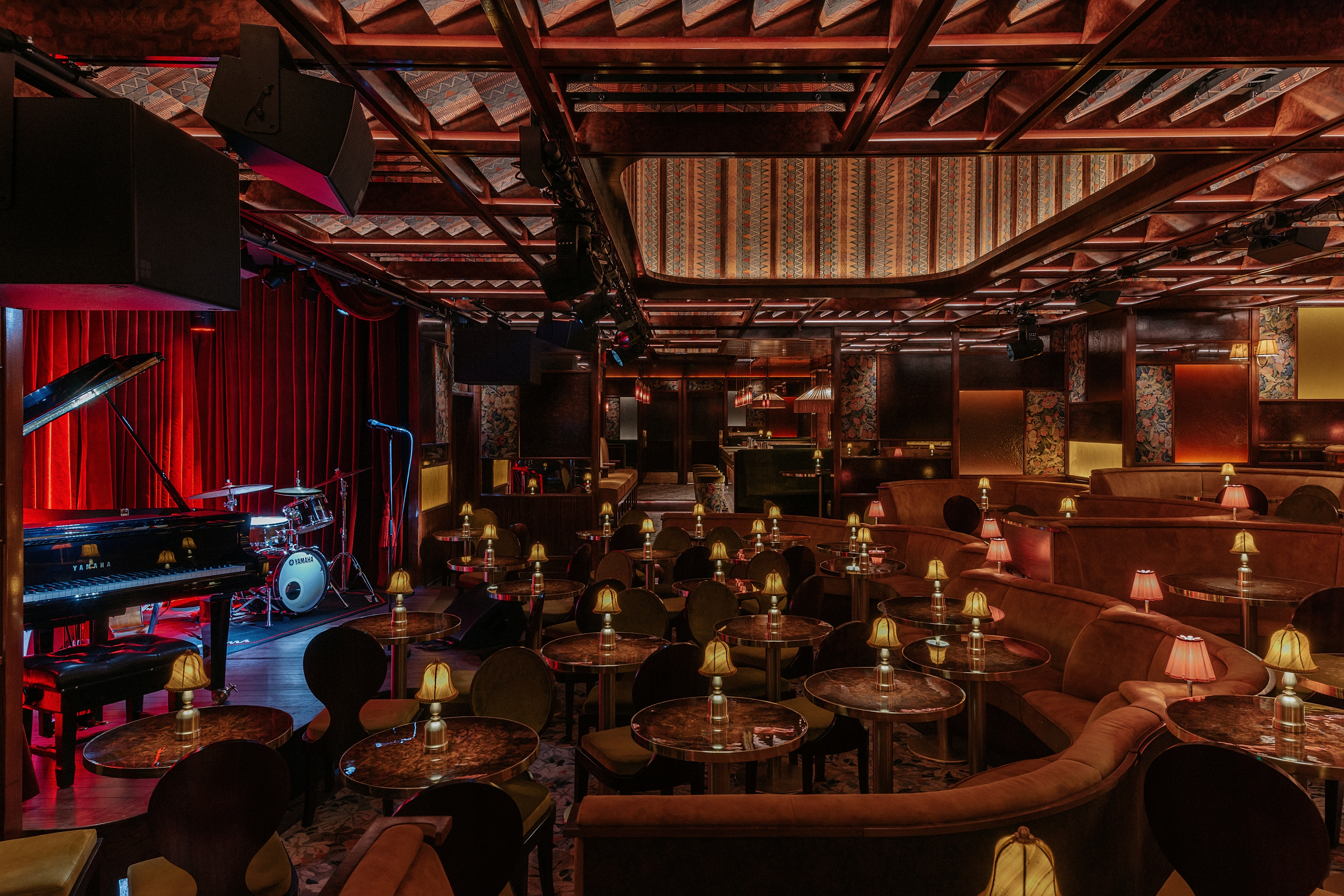 A dimly lit, textiles and lacquered wood-clad jazz club with lots of softly glowing table lamps, performers playing live, and a red-tinted light.