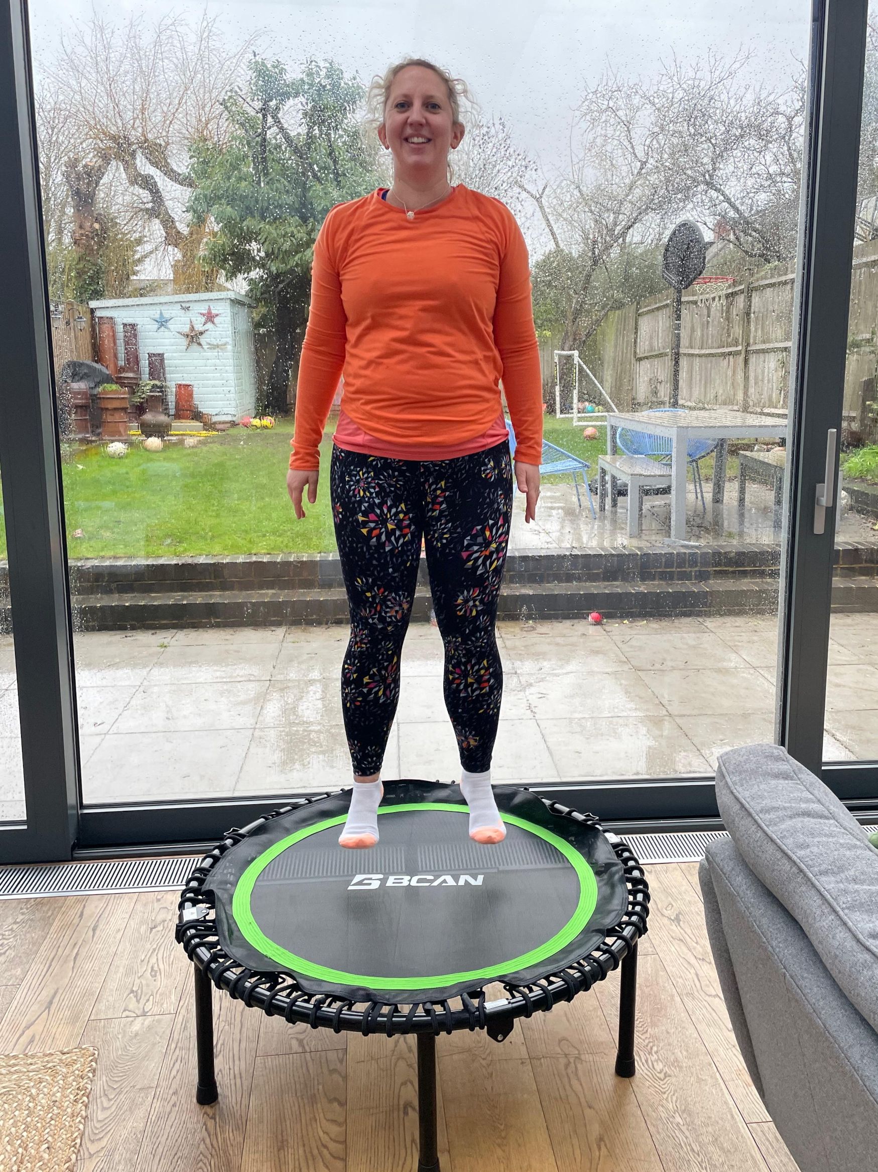 Woman bouncing on small trampoline in domestic setting, a view of a small garden is seen through patio doors